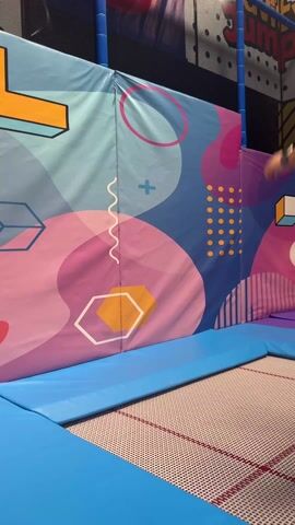 A man is performing gymnastics on a trampoline. He is wearing a pink t-shirt, khaki pants, and bright orange socks. The trampoline has blue padding around it. Behind the man is a colorful wall with geometric shapes and the word "Jump" visible at the top. The man does a backward roll, bounces off the wall, and lands back on the trampoline.