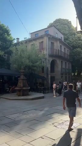 The video pans across an outdoor seating area of a restaurant or cafe situated on a cobblestone plaza. A large, ornate stone fountain stands in the foreground, slightly off-center to the left. To the left of the fountain, under a black umbrella, tables and chairs are occupied by patrons. Behind the tables, a building with large windows is visible. A sign hanging from the building reads "hotel-restaurante RUAS". Further back, more tables and people are visible, with trees providing shade. The scene is bathed in sunlight, suggesting a warm, daytime setting. Several people are walking in the background, and a few are seated at tables. The overall impression is one of a lively, outdoor dining or social gathering space.