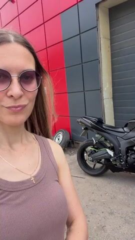 A woman with long brown hair and sunglasses is in front of a building with a red and gray facade. A black motorcycle is parked behind her to the right. The woman is wearing a sleeveless pink top and a gold cross necklace. She makes a kissing face at the camera, then smiles. The video is filmed outdoors in what appears to be a commercial or industrial area.