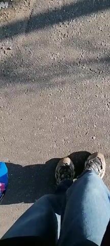 The video begins with a close-up view of a person walking on a paved surface. The person is wearing blue jeans and a light-colored sneaker. The camera appears to be held by the person who is walking. The perspective is that of the person taking the steps. As the person walks forward, the camera angle changes to show a wider view of the environment. A paved road leads to a slight incline with concrete steps. There are trees and greenery along the sides of the road. The sky is blue with some clouds. A metal structure can be seen in the background, possibly a railway or utility tower.