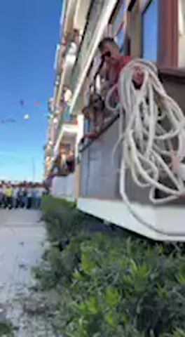 The video captures a daytime scene on a sunny day in Vejer de la Frontera, Spain. A person on a first-floor balcony of a building is seen lowering a thick white rope towards the ground. Other individuals are also visible on balconies, observing the activity.

Below the balcony, at street level, there are green bushes along the building's facade. A dark, animal-like shape, possibly a bull effigy or a real bull, is partially visible lying within these bushes. In the background, a blurred crowd of people can be seen gathered on the street, and a string of colorful festive flags hangs across the street. The sky is clear and blue.

The audio features a voice speaking Spanish, stating "Bueno, ya estamos comprobando que ya se van a proceder con las labores," which translates to "Well, we are already checking that the tasks are about to begin," indicating the start of an event or activity.