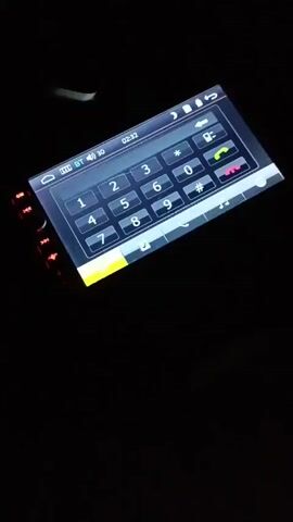 The video shows a close-up, tilted view of a car's digital display screen in a dark environment. The screen displays a virtual keypad for making phone calls, with numbers 0-9, * and #. At the top of the screen, there are icons indicating Bluetooth connection, battery level, and the current time, which reads 02:32. There are also call icons for answering and ending calls. To the left of the screen, there are physical buttons with colored LED lights, which are illuminated in red and then turn green during the video. The lighting in the scene is minimal, with the primary illumination coming from the car's display screen itself. There are no people visible in the video.