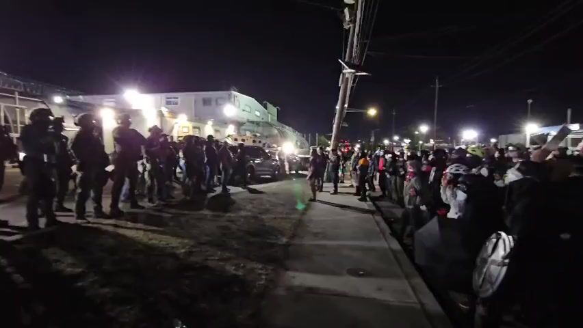 At night, a line of law enforcement officers in tactical gear faces a crowd of protestors across a street. The officers are standing on a grassy area, while the protesters are on a sidewalk. Streetlights illuminate the scene, and a large white building with lights is visible in the background behind the officers. A vehicle is parked between the officers and the building. Some protesters hold signs, and one person is holding a shield. The protesters are chanting, "ICE, KKK, whole thing gotta go." The overall atmosphere seems tense, with a clear division between the authorities and the demonstrating group.