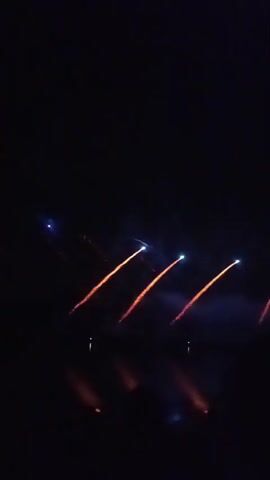 The video shows a nighttime fireworks display over water. Several fireworks are launched in succession, leaving trails of orange sparks and blue lights as they ascend. The reflections of the fireworks are visible on the dark water below. The sky is completely dark, indicating it is nighttime. There are no people or specific objects clearly visible, other than the fireworks themselves and their reflections.