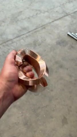 The video shows a close-up of a person's hand holding a coiled piece of copper. The copper appears to be a heat sink, possibly from a computer or other electronic device, and it's bent into a somewhat irregular, looped shape. The hand is gripping the object, and the fingers are visible, along with the textured skin of the palm. The copper itself has a bright, metallic sheen and is made up of many thin, parallel fins.

The background is a concrete floor, suggesting an indoor or possibly an outdoor setting with a paved surface. There are some faint light reflections on the concrete, and in the upper right corner of the frame, a few metallic objects, possibly tools or more scrap metal, are partially visible. The lighting is relatively bright, indicating daytime.

The person is actively manipulating the copper object, causing it to blur slightly with movement, suggesting they are turning it or examining it. There is no discernible emotion conveyed by the hand or the limited view of the person. The focus is entirely on the copper object and the hand holding it. The setting appears utilitarian and industrial or perhaps a workshop environment. No text is visible in the video.