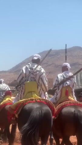 The video shows a procession of riders on horseback, seen from behind. The riders are dressed in traditional striped robes and headscarves, and carry long poles. The horses are adorned with colorful saddle blankets. In the background, there is a mountain range and some buildings. The sky is clear and blue. The scene appears to be outdoors, possibly during a festival or ceremony.