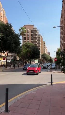 The video shows a street scene in a city on a sunny day. A red car is driving towards the camera on the right side of the street. There are buildings on both sides of the street, and trees are planted along the sidewalks. Several other cars are visible in the distance, driving in the same direction. The sky is blue with a few white clouds. There is a traffic light visible in the distance. The camera is stationary and the video is a single shot.