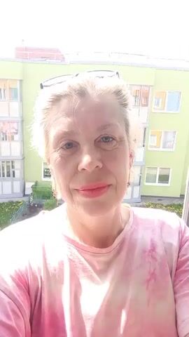 A woman with blonde hair, wearing a pink t-shirt and glasses on her head, is smiling at the camera. She is standing on a balcony. Behind her is a multi-story building painted in light green and white. There is a garden visible below the balcony. The light is bright and appears to be sunny.