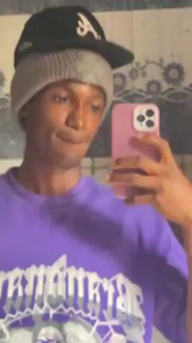 The video shows a young man taking a selfie in front of a mirror. He is wearing a purple t-shirt with a white graphic design, a gray beanie, and a black baseball cap. He is holding a pink smartphone in his right hand, presumably to take the selfie. The background appears to be a tiled wall, possibly in a bathroom. The man is looking at the camera with a slightly quizzical expression.