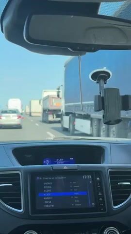 The video shows the view from the driver's seat of a car on a highway. The car is stopped in traffic, with several trucks and cars ahead. The radio display shows that the time is 17:33 and a station called "Radio 1" is playing. The sky is blue and clear, suggesting it is daytime.