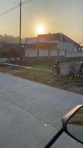 The video shows a first-person perspective of someone riding a motorcycle at sunrise. The sun is low in the sky, casting a warm glow over a rural landscape. The rider is on a paved road, and the side mirror of the motorcycle reflects the scene. In the background, there are houses, trees, and fields. The overall mood is peaceful and serene.