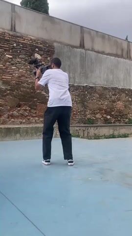 A man in a white t-shirt and dark pants is filming a skateboarder performing a trick. The skateboarder is in the process of landing on the blue court after a jump. The court is surrounded by a textured wall and a concrete structure. The sky is overcast.