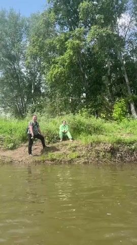The video shows a group of people on a river on a sunny day. The first frame shows two people standing on the bank of the river. The man is on the left, wearing a black t-shirt, black pants, and a black backpack. The woman is on the right, wearing a light green tracksuit. The second and third frames show the perspective from a boat on the river, looking towards the bank with lush green trees and vegetation. The fourth frame shows two inflatable boats on the river. The boat in the foreground is partially covered with a grey tarp and has a black life vest on it. In the background, in another boat, are two people wearing life vests, one yellow and one orange. They are facing away from the camera. The water appears calm and slightly murky. The sky is blue with scattered white clouds.