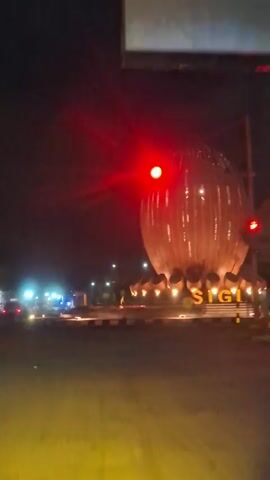 A nighttime video shows a large, illuminated sculpture with vertical lines and rounded edges, possibly representing a flower or fruit. The sculpture is set against a dark sky. In the foreground, a traffic light is visible, with the red light illuminated. The sculpture is labeled with the word "SIGI" in large yellow letters. The video pans slightly to the right, revealing a street with some lights and moving vehicles in the distance. The overall scene is dark, with the sculpture and streetlights providing the primary illumination.