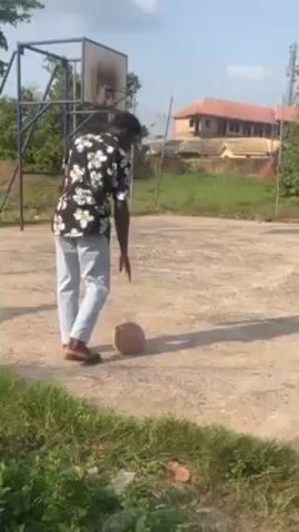 A young man is walking on a basketball court while holding a basketball. The court is outdoors, on a sunny day. The man is wearing a black and white floral shirt, light-colored pants, and sandals. There is a basketball hoop and net behind him. Other people are in the background. The man is looking down at the basketball as he walks.