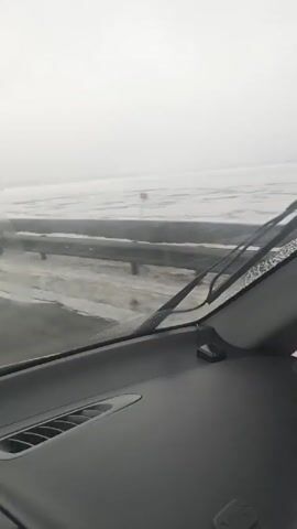 The video shows a view from the dashboard of a car driving on a highway in snowy, foggy conditions.  A metal guardrail runs along the right side of the road.  Beyond the guardrail, a flat, icy landscape stretches out to the horizon.  In the car's side mirror, the headlights of another vehicle can be seen approaching from behind. The windshield wipers are active, clearing moisture from the glass.  The overall scene appears cold and bleak.