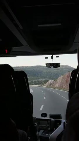 The view from inside a bus looking forward shows a gray asphalt road curving ahead through a hilly, green landscape. The bus interior is visible, including the backs of the seats, the rearview mirror, and the driver's console. The sky is overcast. A small display shows the number seven, and another shows the time as 11:27.