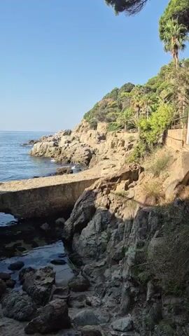 A narrow concrete walkway extends from a rocky cliffside over a small body of water. The water is clear, with rocks visible beneath the surface. The walkway is partially submerged at its end. The cliffside is covered in rocks and vegetation, including palm trees and other greenery. The sky is clear and blue. The scene appears to be a coastal landscape.