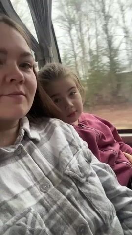 A woman wearing a grey and white plaid shirt sits next to a young girl wearing a red sweatshirt in what appears to be a moving vehicle. The girl has her head resting on the woman's shoulder. A window is visible behind them, showing trees passing by.