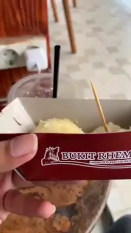 Here is a concise description of the video:

The video shows a person holding a small, maroon-colored cardboard container with the logo of "Bukit Rhema: House of Prayer for All Nations" printed on it. Inside the container, there appear to be two portions of food, possibly dessert, with a toothpick stuck into one of them. The camera then focuses on the container against the backdrop of a balcony with a view of a green, hilly landscape. The sky appears overcast.