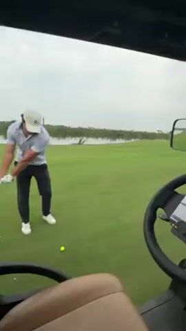 From inside a golf cart, a man wearing a white cap, gray shirt, and dark pants is seen swinging a golf club. The golf ball is yellow and sits on the green grass in front of him. A pond or lake is visible in the background with greenery along the banks. The golf cart's dashboard is visible, including the steering wheel, a scorecard, and side mirror. After the swing, the camera view pans forward, following the ball's trajectory down the fairway.