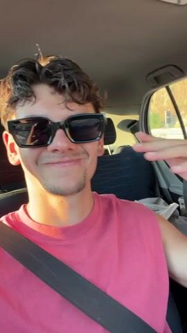 The video shows two people in a car. In the foreground, a young man wearing sunglasses and a pink sleeveless shirt smiles at the camera and gestures with his hand. He is wearing a seatbelt. In the background, a young woman wearing sunglasses and a white tank top and pink pants is driving. She is also wearing a seatbelt. The car appears to be moving on a road with foliage visible outside the windows. The lighting suggests it is daytime.