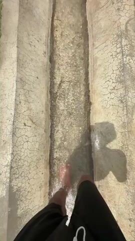 The video shows a person's bare feet walking through a shallow concrete channel filled with flowing water. The channel appears to be outdoors, with dry, cracked concrete on the sides and grass on the edges. As the camera moves forward, other people become visible. One person is standing on the left side of the channel, and two people are walking on the right side, one wearing brown shoes. Further ahead, a person in yellow shorts walks alongside the channel, and a child runs down the channel in the water. Trees and a red object, possibly a bicycle, are visible in the background. The overall impression is of a casual outdoor activity, possibly a way to cool off or play in the water.