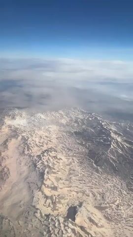 The video captures an aerial view from an airplane, showcasing a vast and rugged mountainous landscape below. The terrain appears arid and is extensively covered with patches of white snow, particularly on higher elevations and within valleys. A clear blue sky is visible above, with some scattered white and grey clouds situated between the aircraft and the ground. From the 0:01 mark, the wing of the airplane, featuring a light color and a distinct pinkish-red wingtip, becomes visible on the right side of the frame, providing context to the aerial perspective. The scene depicts a bright, sunny day during a flight over what is identified as the region of Gündoğmuş, Turkey. No people or visible text are present in the video.