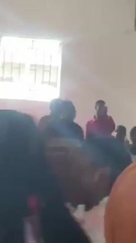 The video shows a classroom scene with numerous people present. Most are seated at desks, facing a person who stands and appears to be addressing them. This individual is wearing traditional African attire, including a patterned shirt and a hat. The students' faces are not clearly visible due to the camera angle, but they appear to be attentive to the speaker. The classroom itself has white walls, a window with bars, and a ceiling fan. There is a blackboard on one side of the room. The atmosphere seems informal and relaxed, with some students smiling.