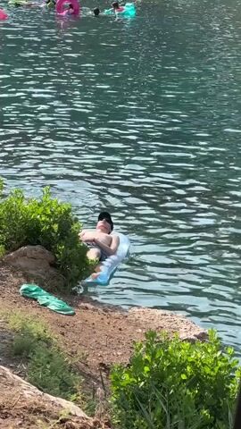 The video shows a person lying on an inflatable float in a body of water. The person is wearing a hat and appears to be relaxing. There are other people and inflatable objects visible in the background, suggesting a recreational setting. The water is calm and the surrounding area appears to be a natural landscape with vegetation and rocks. The lighting suggests it is daytime.