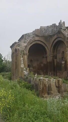The video shows a close-up, exterior view of a ruined stone church. The church has arched openings and a domed roof topped with a cross. The stone structure is weathered and shows signs of significant decay, with some walls crumbling and vegetation growing around it. The surrounding environment appears overgrown with tall green grass and some small yellow flowers. The sky is overcast, suggesting a cloudy or foggy day. No people are visible in the video. The camera slowly pans across the front and side of the ruined structure.