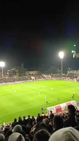 The video shows a soccer match being played at night in a stadium with bright floodlights. The field is green, and there are players in blue and red uniforms. The stands are full of spectators, most of whom are dressed in dark clothing, with some wearing hats or hoodies. Many fans are gathered in a section near the goal post, waving red and white flags. In the background, buildings of varying heights can be seen. The sky is dark and clear. The camera is positioned in the stands, providing a view of the field and the crowd. The crowd is cheering and chanting.