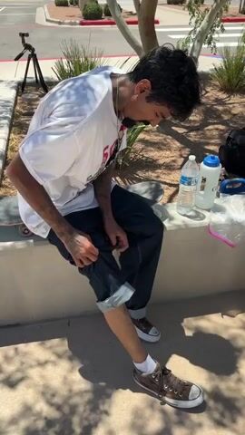 A person is sitting on a concrete ledge outdoors. They are wearing a white t-shirt, dark jeans with cuffed legs, white socks, and brown shoes. The person is looking down at their right knee, which has a scrape with some redness and possibly blood. There is a tattoo of a spiderweb and spider on the person's lower leg. A skateboard is visible behind them, and in the background, there is a tripod with a camera, a water bottle, a larger water jug, and a plastic bag. The ground is paved, and there are trees and bushes in the background. The lighting suggests it is daytime.