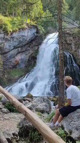 A young person wearing a white t-shirt and black shorts sits on a rock, facing away from the camera, and looking at a large waterfall cascading down a rocky cliff face. The waterfall is surrounded by lush green trees and vegetation. In the foreground, a wooden fence runs horizontally across the frame, with a large fallen log resting on it. Another person, partially visible and wearing a blue shirt, walks along a path to the left of the waterfall. The ground is covered with rocks and dirt, and the overall environment appears to be a natural, wooded area.