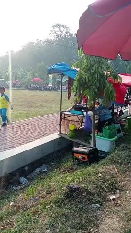 A young boy in a yellow and blue shirt walks along a brick path. Behind him, there are several market stalls set up outdoors under awnings. One stall has a sign that reads "DEGAN, ES DE & DEGU WU". A man in a green shirt is working at this stall, which is set up on the back of a pickup truck filled with coconuts. Other items visible include coolers, a generator, and various supplies. The ground is covered in grass and some debris. In the background, more people are gathered, and there are trees and vegetation. The sun appears to be low in the sky, suggesting it is either early morning or late afternoon.