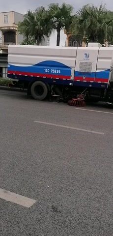 A street sweeper truck, predominantly white with blue and red wavy stripes, is operating slowly on a paved road in Ha Tu Ward, Vietnam. The truck has the license plate "14C-259.94" and visible rotating brushes for cleaning the road. On its side, the brand "THUY DUONG" is visible. As the sweeper moves, a person on a motorcycle passes by, carrying a large blue object on the back. Shortly after, a red bus with white stripes, featuring the partial text "mobilhome," also drives past. The scene is set during the daytime, with palm trees and buildings lining the street, and mountains visible in the background under an overcast sky.