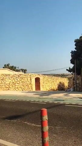 The video pans to the right, showing a stone wall against a clear blue sky. A paved road with a green lane is visible in the foreground, bordered by red and white striped bollards. The stone wall is constructed of large, rough-hewn blocks. Further along, the wall transitions to a section made of smaller, more irregularly shaped stones. A doorway with a dark red door is set into the wall, topped with a small niche containing a statue. The wall continues, with a dark window visible, and further on, a green door and a building with a tiled roof. Overhead wires and a cell tower are also visible. The scene appears to be a street or roadside in a sunny, possibly Mediterranean, location.