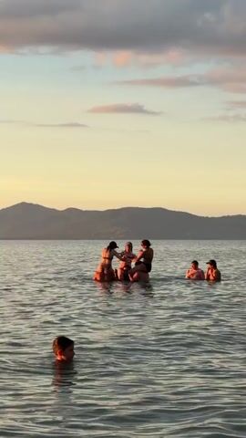 A group of people are in the ocean. In the foreground, a person is visible from the shoulders up, facing towards the right and reaching their left arm forward. In the midground, several people are gathered in the water. Two people appear to be sitting on the shoulders of a third person. Two other people are in the water to the right of this group, with their heads and shoulders visible. The sun is setting, casting a warm glow on the water and the sky, which has clouds. Mountains are visible in the background.