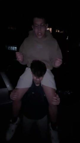Two young men are outdoors at night, one giving the other a piggyback ride. The person being carried wears a tan hoodie, shorts, and sneakers. He also wears glasses. He holds onto the other man’s head, who wears a dark shirt, gray pants, and sneakers. The person being carried raises his hands up in the air in different positions. A car is visible in the background.