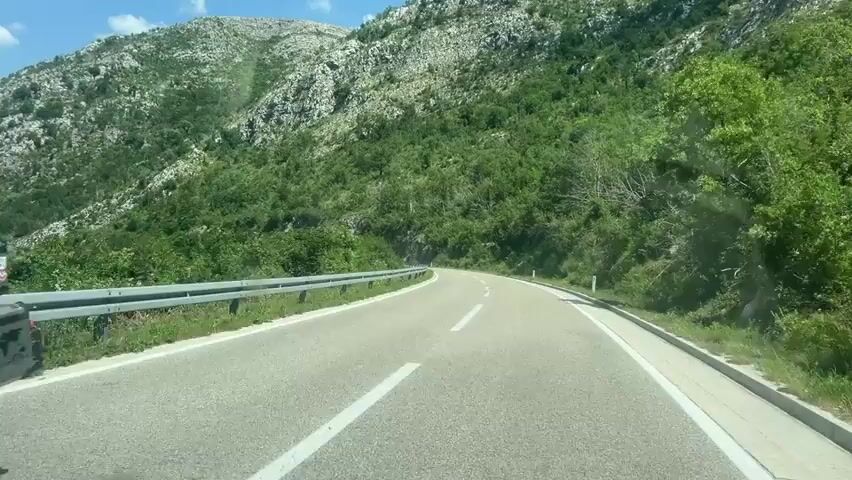 The video shows a view from a moving vehicle on a paved road. The road is curved and has white lane markings. A metal guardrail runs along the left side of the road. The environment is mountainous and green, with lush vegetation and rocky slopes visible. The sky is blue with some scattered white clouds. The perspective suggests the camera is positioned on the dashboard or windshield of a car. The lighting indicates it is daytime, likely sunny. There are no people or other vehicles directly visible in the frame. The road appears to be in a rural or natural setting.