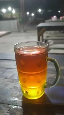 This is a close-up video of a glass mug filled with a dark amber liquid, likely iced tea, sitting on a wooden table. The mug has a textured pattern of circular indentations and a green handle. The lower portion of the mug is illuminated by a yellow light source, casting a warm glow. The background is blurred, showing an outdoor setting at night with lights from buildings or lamps visible in the distance. There are also hints of tables and chairs, suggesting a restaurant or outdoor seating area. The focus is entirely on the beverage.