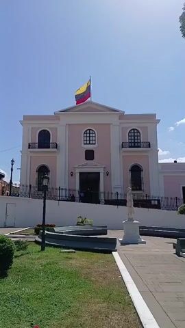 The video shows a two-story building with a pink facade and white trim. The Venezuelan flag is flying on top of the building. There are three arched windows on the second floor with small balconies. On the ground floor, there is a central doorway, flanked by two arched windows. A black wrought-iron fence surrounds the property. In front of the building is a small park with a statue, benches, and a street lamp. The sky is blue with some clouds.  The video appears to be taken from a stationary position.