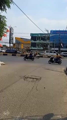 A sunny day shows a street intersection with several motorcycles driving. The road has visible cracks and potholes. Buildings line the street, some appearing to be commercial establishments with signs. Red and white flags are mounted on poles along the road. The sky is clear and blue.