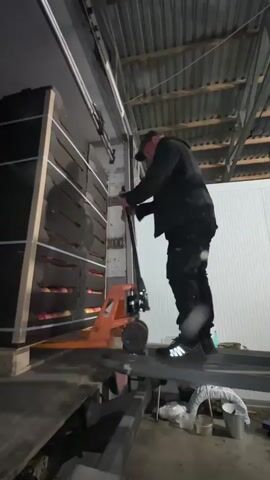 A man in black clothing and a baseball cap is operating an orange pallet jack. He is standing on a metal platform, holding the handle of the pallet jack and pulling it towards him. To the left of the man is a tall, dark shelving unit filled with what appear to be rows of apples. The shelving unit is leaning against a wall. The man is positioned at the end of the metal platform, which appears to be a loading dock or ramp. The ceiling above is corrugated metal. The lighting is dim.