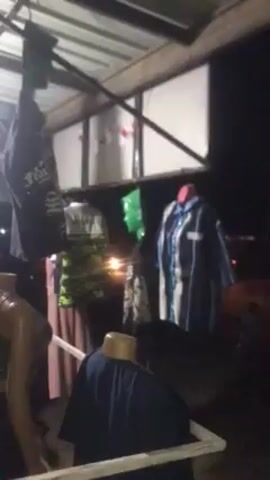 The video shows a dark outdoor setting, likely at night. Several mannequins are displayed under a covered area, showcasing various clothing items. A row of square panels lines the back wall of the covered space. A light source, possibly a vehicle's headlights, shines from a distant road, illuminating the scene intermittently. The camera is relatively still but the focus may be shifting. The scene appears to be a small roadside clothing display or market.