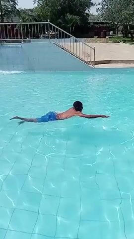 Here is a description of the video:

The video shows a man swimming in a pool. He uses an overhand stroke. The pool has a light blue tile bottom, and the water is clear and a lighter blue. The pool is surrounded by a tiled deck area with a metal railing. Beyond the deck are trees, shrubs, and covered seating areas. The video appears to be taken during daylight.