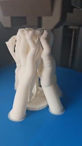 This is a close-up, slightly shaky, indoor video of a 3D printer actively printing a white object. The object appears to be a cluster of abstract, elongated, vaguely humanoid shapes that are connected at the top and flare out at the bottom like legs or columns. The printer's nozzle is visible at the top of the frame, just above the printed object, suggesting it is in the process of adding material. The object rests on a blue print bed. The background is dark and blurry, suggesting the focus is solely on the printing process.