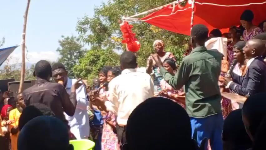 A group of people are gathered outdoors under a red and blue makeshift canopy. Many are singing into microphones, while others play instruments like guitar and drums. The group is diverse, with both men and women dressed in various outfits. Some individuals are dancing and clapping along with the music. The crowd seems to be enjoying the performance, and the atmosphere is lively and celebratory. Trees and bushes can be seen in the background, suggesting the event is taking place in a garden or park.