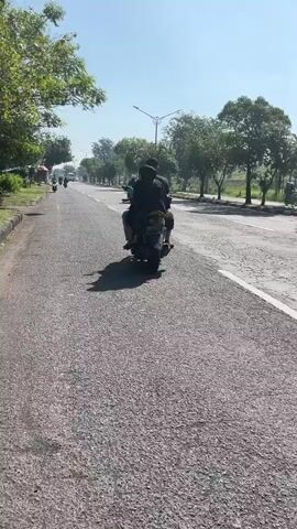 The media shows a road scene with two people riding a scooter. The subjects are two individuals, their faces obscured from view as they ride away from the camera. They are seated on a dark-colored scooter, with one person in front and the other behind.

The scene is a paved road, appearing to be a two-lane highway, with a clear sky overhead. Lush green trees line the sides of the road, and in the distance, more trees and vegetation are visible, suggesting a rural or semi-rural setting. A single lamppost stands in the middle distance. The road surface is asphalt, showing some wear and tear with visible cracks. There are white painted lines demarcating the lanes.

The activity is the act of traveling on a scooter. The time of day appears to be morning or early afternoon, indicated by the bright sunlight casting shadows. The weather seems to be clear and sunny. The location context is Kedungbendo, Indonesia.

Notable details include the presence of other vehicles further down the road, also appearing to be motorcycles, suggesting active road use. The overall mood is one of movement and daily life in a tropical or subtropical environment. No visible text is present in the media file.