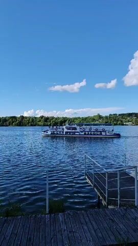 The video captures a tranquil scene on a wide, blue lake under a bright, sunny sky with scattered white clouds. A medium-sized, white passenger boat with blue accents cruises across the water, its wake creating ripples. The boat appears to have multiple decks and numerous people visible on its upper level, suggesting a leisurely excursion or ferry service.

In the foreground, a weathered wooden dock with metal railings juts out into the water, indicating a potential boarding or viewing area. The shore opposite the boat is lined with a dense forest of green trees, with glimpses of what might be buildings or structures peeking through the foliage in the distance, hinting at a populated shoreline. The overall impression is one of a calm, pleasant day, likely in the late morning or early afternoon, given the bright sunlight and clear sky. The setting evokes a sense of peace and natural beauty, typical of a summer day by the water. No visible text is present in the video.